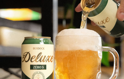Green can of Burdock Deluxe Zero 5 non-alcoholic beer being poured into a glass with a blurred background