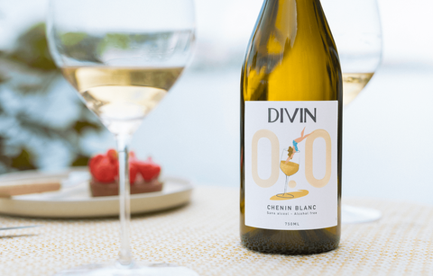A golden bottle of DIVIN Non-Alcoholic Chenin Blanc wine on a table with a glass of wine and fruit in the background.