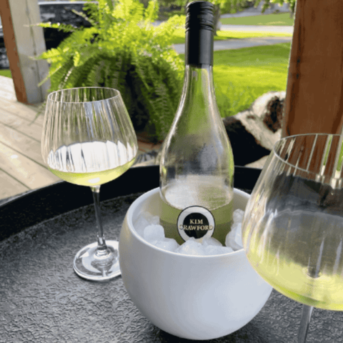 Bottle of white wine in an ice bucket with two glasses on a table outdoors.