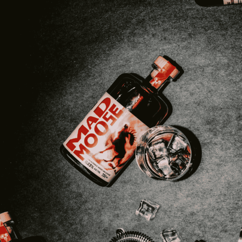 Bottle of Mad Moose Non-Alcoholic Whiskey beside a glass filled with ice, on a dark chalk background