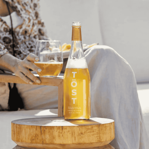 Bottle of Töst non-alcoholic sparkling tea on a wooden table with a person holding a glass in the background.