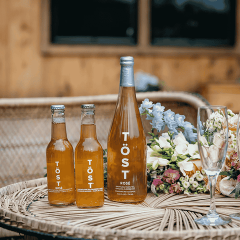 Three bottles of TÖST Rosé wine on a wicker table with flowers and a glass.