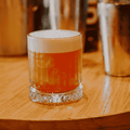 A lowball cocktail with a thick layer of white foam sits on a round wooden bar table, with metal shakers blurred in the background.