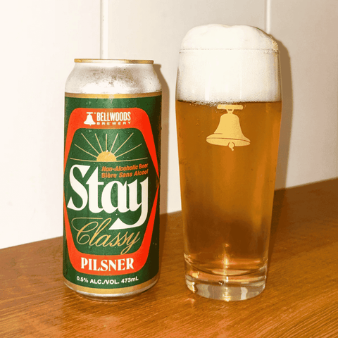 A can of Bellwood's Brewery Stay Classy non-alcoholic Pilsner beer stands next to a tall glass of the golden beer with white foam, set on a wooden table against a plain wall.