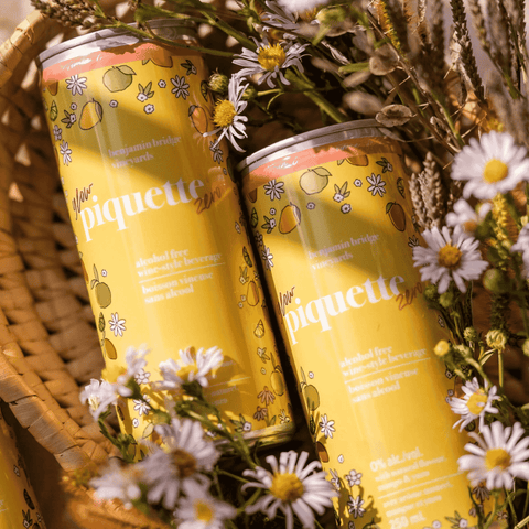 Two yellow cans of Piquette Zero Glow by Benjamin Bridge with floral decorations on a woven basket and flowers background.