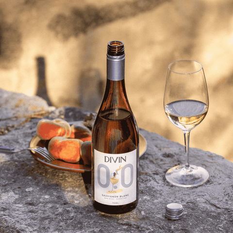Bottle of DIVIN 0.0 Sauvignon Blanc with a glass of wine on a stone surface with a plate of peaches