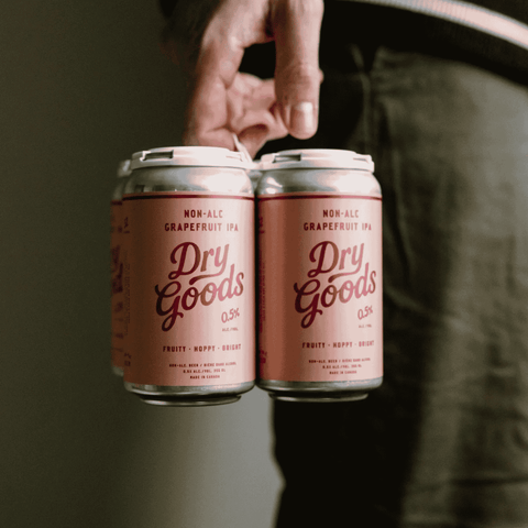 A person holding a 4-pack of Dry Goods Non-Alcoholic Grapefruit IPA