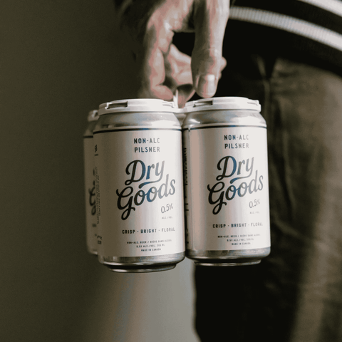 A person holding a 4-pack of Dry Goods Non-Alcoholic Pilsner