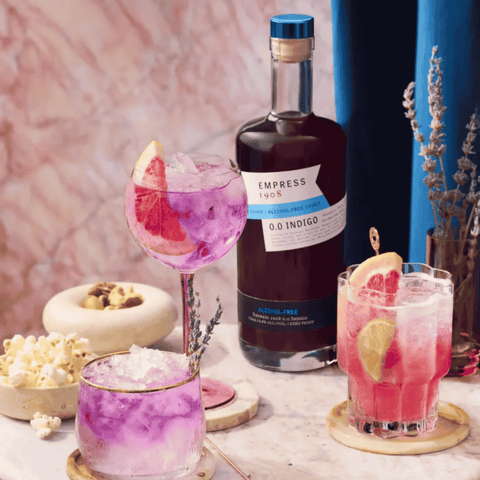 Bottle of Empress 1908 O.O. Indigo gin with pink cocktails on a marble table.