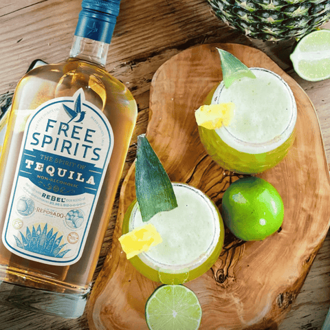 A bottle of Free Spirits non-alcoholic Tequila on a wood table with limes, pineapples, and a non-alcoholic cocktail