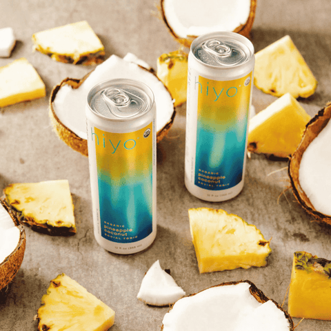 Two cans of hiyo Organic Pineapple Coconut are arranged on a textured gray surface, surrounded by fresh pineapple slices and halved coconuts.