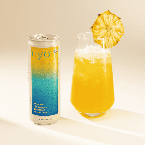 A can of Hiyo Pineapple Coconut Social Tonic poured into a glass with ice and garnished with a dried pineapple slice