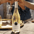 A bottle of Kim Crawford Illuminate Alcohol-Removed Sauvignon Blanc on a table with people cheersing glasses in the background