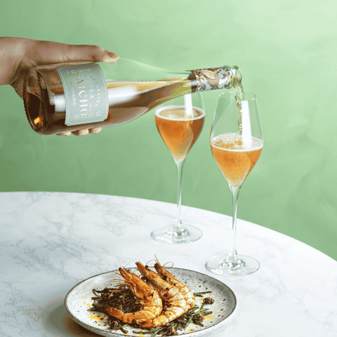 A bottle of Saicho Sparkling Hojicha Tea being poured into two glasses paired with a prawn dish