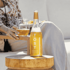 Bottle of Töst non-alcoholic sparkling tea on a wooden table with a person holding a glass in the background.